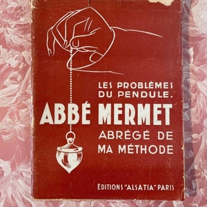 May include: A red book cover with a white illustration of a hand holding a pendulum. The book title is "Les Problèmes du Pendule. Abbé Mermet Abrégé de Ma Méthode" and the publisher is "Editions "Alsatia" Paris".