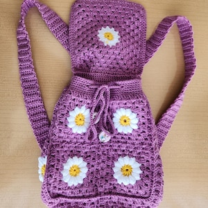 May include: Purple crocheted backpack with a daisy pattern and a drawstring closure. The backpack has two adjustable straps.