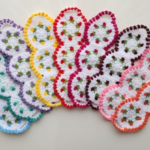 Può includere: Un assortimento arcobaleno di presine all'uncinetto in vari colori, tra cui blu, verde, viola, giallo, rosso, rosa e arancione. Ogni presina presenta un centro bianco con un motivo floreale verde.