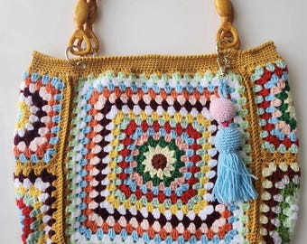 May include: A handmade crochet tote bag with a wooden handle. The bag features a colorful granny square design in shades of red, yellow, blue, and green. A blue tassel and pink pom-pom hang from the handle.