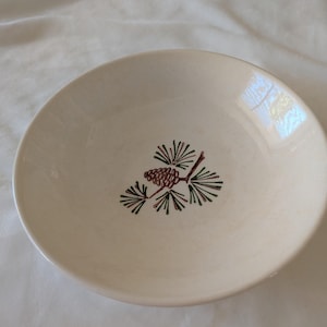 Peut inclure: Bol en céramique blanc cassé avec un motif de branche de pin. La branche présente une pomme de pin marron et des aiguilles vertes. Le bol est rond et possède un bord légèrement surélevé. Design simple et élégant pour servir des aliments.