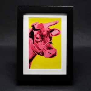 Puede incluir: Una impresión enmarcada de la cabeza de una vaca rosa sobre un fondo amarillo.