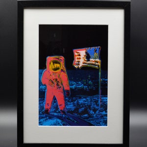 Puede incluir: Una impresión enmarcada de un astronauta de pie en la luna con una bandera estadounidense plantada en la superficie lunar. El astronaute lleva un traje espacial rojo y amarillo y la bandera ondea al viento. El fondo es un cielo azul oscuro y negro con estrellas.