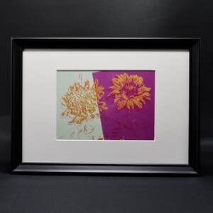 Puede incluir: Una impresión enmarcada de una ilustración abstracta de flores de colores. La flor es amarilla y naranja con un fondo rosa. La impresión está enmarcada en un marco negro.