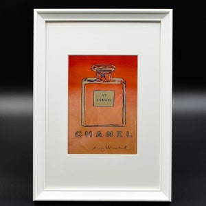 Puede incluir: Obra de arte enmarcada con una ilustración de un frasco de perfume. La imagen tiene un fondo degradado cálido en naranja y dorado. La botella está etiquetada "N°5 CHANEL" con el nombre de la marca y la firma del artista debajo.