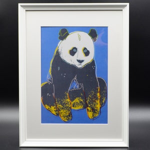Puede incluir: Una impresión enmarcada de un oso panda blanco y negro sobre un fondo azul. El panda está sentado con sus patas delanteras frente a él. La impresión está en un marco blanco.