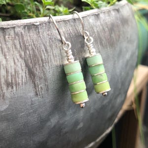 May include: A pair of dangle earrings featuring stacked cylindrical light green beads separated by thin silver-toned metal spacers. The beads are suspended from silver-toned metal ear wires, creating a vertical, layered design with a smooth finish.