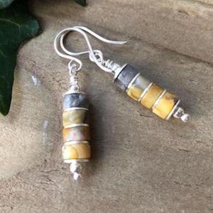 May include: A pair of dangle earrings with silver-tone hooks. Each earring features a stack of cylindrical beads in shades of gray, yellow, and brown. The beads are separated by silver-tone accents. The earrings are displayed on a wooden surface.