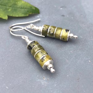 May include: A pair of dangle earrings featuring three cylindrical olive green stone beads with dark speckles, separated by thin silver-toned metal spacers. The earrings are finished with a silver-toned wire hook and a small spherical bead at the base.