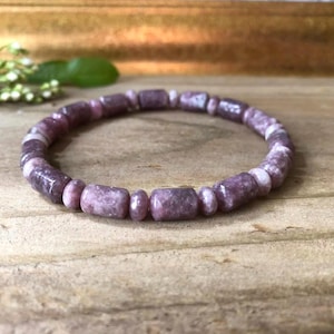 May include: A beaded bracelet featuring a series of small, rectangular and round, light purple beads. The bracelet is displayed on a wooden surface, with a blurred background.