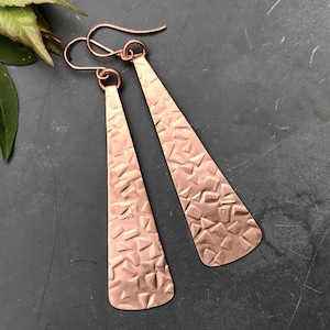 May include: A pair of long, dangling copper earrings featuring a flat, elongated triangular shape. The surface of each earring is textured with a repeating pattern of small, irregular geometric indentations and hangs from a simple wire hook.