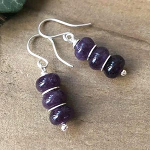 May include: A pair of dangle earrings featuring three deep purple beads separated by silver-colored rings. The earrings have silver-colored hooks and small silver-colored beads at the bottom. The earrings are displayed on a light brown surface.