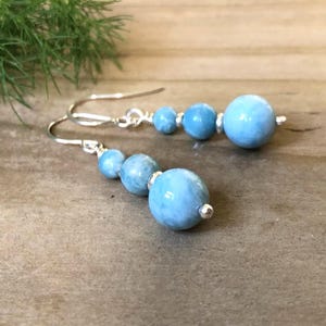 May include: Dangle earrings with light blue spherical beads of different sizes. The beads are strung on silver-coloured hooks and connectors. The earrings are presented on a textured, neutral-toned surface.