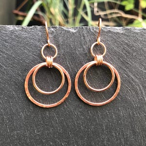 May include: A pair of copper-coloured hoop earrings. Each earring has two connected circles, one larger than the other. The earrings hang from a small hook. The jewellery is displayed on a dark grey surface.