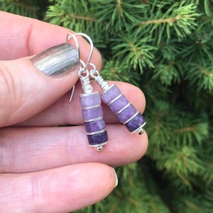 May include: A pair of drop earrings featuring a vertical stack of four cylindrical beads in an ombre purple colour gradient, ranging from light lavender to deep violet. The beads are separated by thin silver-toned metal spacers and hang from silver-toned ear wires.