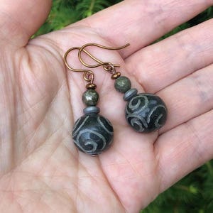 May include: A pair of handcrafted earrings featuring dark green, patterned beads with copper-colored ear wires. The earrings have a unique design with a combination of round and cylindrical beads, creating an elegant and artistic look. The earrings are displayed against a natural background.