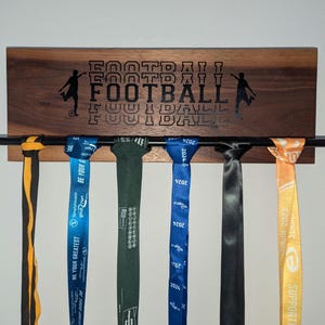May include: A dark wood medal display with the word "FOOTBALL" engraved on it. Five medals are hanging on the display, including a blue, green, black, and orange medal.