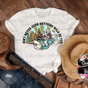 May include: White t-shirt with a graphic design of a skull with a guitar, playing cards, dice, and a bottle of whiskey. The text on the shirt reads "Ain't Never Seen Anything Wild As You".