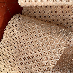 May include: Close-up of several rolls of natural-colored cane webbing. The webbing has a woven pattern of small, evenly spaced holes. The rolls are partially unrolled, revealing the intricate design and texture of the material.