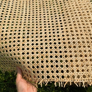 May include: Close-up of a sheet of natural woven cane webbing. The webbing has a repeating pattern of small, evenly spaced holes. The material is a light tan color and appears to be flexible. The edges of the webbing are slightly frayed.