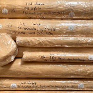 May include: A stack of rolled packages wrapped in brown paper and clear plastic. Handwritten addresses are visible on the paper, with names and street addresses. Small white stickers with handwritten text are also present.