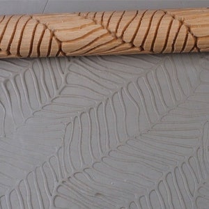 Könnte beinhalten: Ein Holz-Nudelholz mit einem geschnitzten Blatt-Muster wird verwendet, um einen Abdruck auf einer grauen Ton-Oberfläche zu erstellen. Der Abdruck ist ein sich wiederholendes Muster von Blättern.