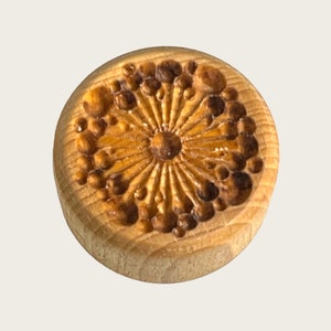 May include: A round wooden stamp with a carved design of a flower with a centre circle and radiating lines. The flower is surrounded by a ring of small circles.