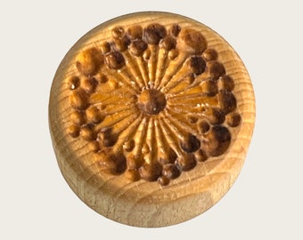 May include: A round wooden stamp with a carved design of a flower with a center circle and radiating lines. The flower is surrounded by a ring of small circles.