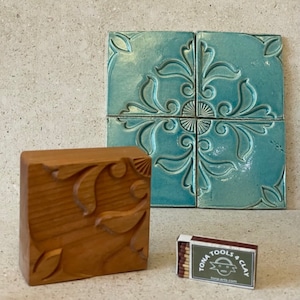 Könnte beinhalten: Eine Holz-Fliesenform mit einem geschnitzten Blumenmuster, neben einem Satz von vier tealblauen Keramikfliesen mit einem ähnlichen Blumenmuster. Eine Streichholzschachtel mit dem Text "TONIA TOOLS & CLAY" befindet sich im Vordergrund.
