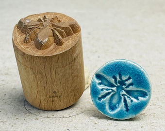 May include: A wooden stamp with a carved bee design and a blue ceramic tile with an impressed bee design.
