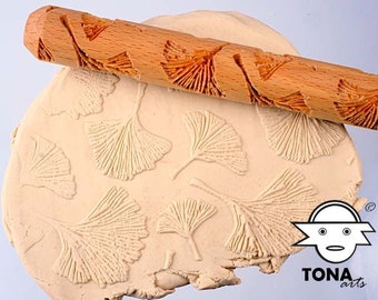 May include: A wooden rolling pin with a carved design of ginkgo leaves. The rolling pin is being used to create an imprint of the leaves in a piece of clay.