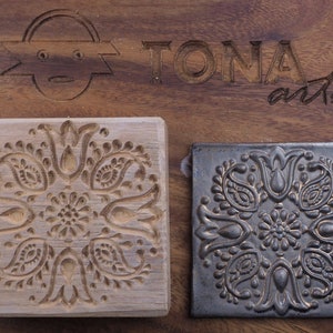 Può includere: Un blocco di legno con un motivo paisley intagliato, accanto a una piastrella nera con lo stesso motivo. La piastrella è di una tonalità di grigio più scura e presenta un motivo più dettagliato. Il blocco di legno è etichettato "TONIA arts".