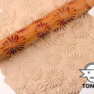 Könnte beinhalten: Ein Holz-Nudelholz mit einem geschnitzten Gänseblümchenmuster wird verwendet, um ein geprägtes Design auf einem Blatt beigefarbenem Ton zu erstellen. Das Nudelholz ist aus Naturholz gefertigt und hat eine glatte, polierte Oberfläche. Der Ton ist weich und formbar, und das Nudelholz hinterlässt einen klaren, detaillierten Abdruck.