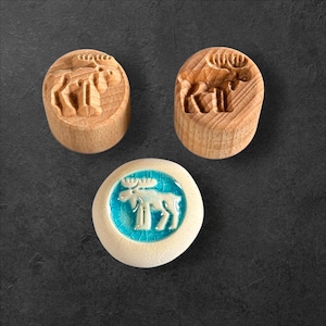 Peut inclure: Trois tampons sur le thème de l'élan. Deux sont cylindriques, sculptés dans du bois, et présentent un motif d'élan. Le troisième est un tampon rond en céramique avec un motif d'élan dans un cercle turquoise.