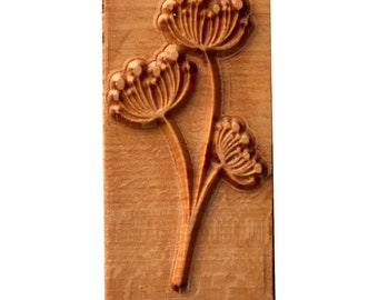 May include: A wooden stamp with a carved design of two stylized flowers with multiple rounded petals. The stamp is made of light brown wood with a natural grain pattern.