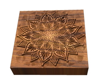 May include: A wooden square block with a carved mandala design. The mandala is intricate and detailed, with a brown color.