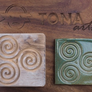 Może przedstawiać: Dwa kwadratowe kafelki, jeden drewniany, a drugi ceramiczny, oba z rzeźbionym wzorem trzech splecionych spiral. Kafel drewniany jest jasnobrązowy, a kafel ceramiczny jest jasnozielony. Kafelki znajdują się na drewnianej powierzchni z wygrawerowanym tekstem "TONA arts".