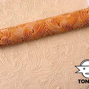 Könnte beinhalten: Ein Holz-Nudelholz mit einem wirbelnden Muster wird verwendet, um ein strukturiertes Design auf einem Stück Ton zu erstellen. Das Nudelholz ist aus Naturholz gefertigt und hat eine glatte, polierte Oberfläche. Der Ton ist hellbraun und hat eine weiche, formbare Textur.