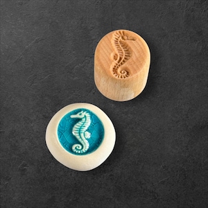 Op de afbeelding: Twee decoratieve items met zeepaardontwerpen. Eén is een houten stempel met een uitgesneden zeepaardje. De andere is een rond, wit keramisch stuk met een turquoise zeepaardontwerp in het midden.