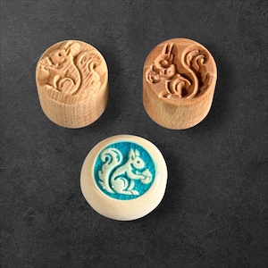Könnte beinhalten: Drei Holzstempel mit Eichhörnchen-Designs. Zwei Stempel sind aus Holz geschnitzt und zeigen ein Eichhörnchen, das eine Nuss hält. Der dritte Stempel hat eine weiße Basis mit einem türkisfarbenen Eichhörnchen-Design.