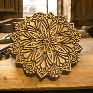 Könnte beinhalten: Ein Holzblock mit detailliertem Blumenmuster. Der Block ist hellbraun mit dunkelbraunen Schnitzereien. Das Design zeigt eine zentrale Blütenform mit strahlenförmigen Blütenblättern und komplizierten Mustern.
