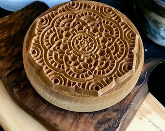 May include: A round, wooden stamp with an intricate, carved mandala design. The stamp is light brown and sits on a dark wooden surface. The detailed pattern features concentric circles and floral elements, perfect for crafting or decorative purposes.