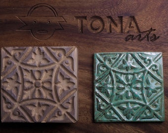 May include: Two square tiles with a decorative pattern. The tile on the left is made of wood and is brown. The tile on the right is made of ceramic and is green.