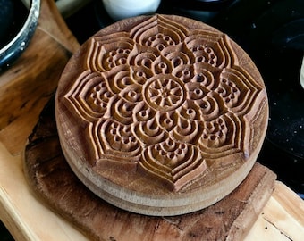 May include: A wooden round stamp with a carved mandala design. The stamp is made of light brown wood and has a detailed pattern of circles, swirls, and leaves.