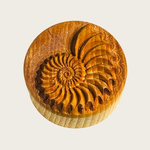 Op de afbeelding: Een houten stempel met een gedetailleerde gravure van een nautilus schelp. De schelp is lichtbruin van kleur en heeft een spiraalvormig patroon.