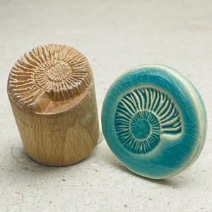 Könnte beinhalten: Ein Holzstempel und ein türkisfarbener Keramikknopf, beide mit einem Ammoniten-Fossil-Design. Der Stempel ist zylindrisch mit einem geschnitzten Spiralmuster. Der Knopf hat eine Craquelé-Glasur und ein erhabenes Fossil-Design.