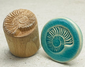 May include: A wooden stamp and a teal ceramic button, both with an ammonite fossil design. The stamp is cylindrical with a carved spiral pattern. The button has a cracked glaze finish and a raised fossil design.