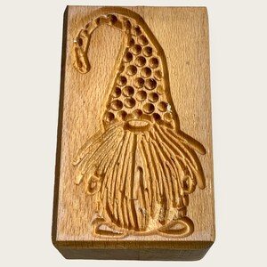 Puede incluir: Cortador de galletas de madera con forma de gnomo con un sombrero puntiagudo y una larga barba. El cortador está hecho de madera marrón claro.