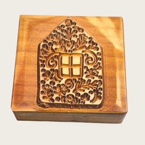 May include: A wooden cookie cutter shaped like a house with a detailed design of flowers and swirls. The house has a window with four panes.