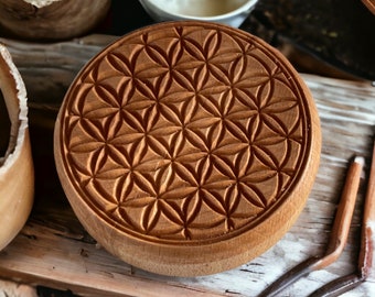 May include: A round wooden box with a carved flower of life design. The box is made of light brown wood and has a natural finish.